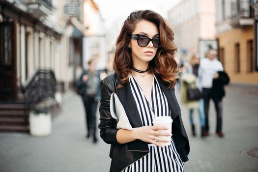 Fashionable brunette woman in sunglasses wearing in striped suite and black leather jacket walking at street and holding cup of coffee. Beautiful and stylish girl with choker on neck, handbag in hand.