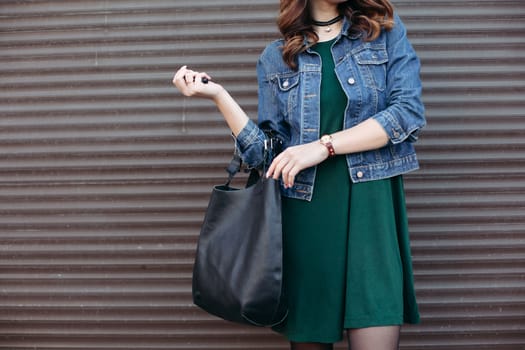 Crop of stylish incognito girl with big black leather bag after shopping, posing at street, leaning on wall. Woman in dress, jeans jacket, with accessories. Street fashion.