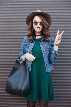 Stylish beautiful brunette woman in sunglasses, green dress, brown hat and big black bag, posing standing at street. Fashionable smiling girl leaning on wall. Concept of fashion and street trend look.