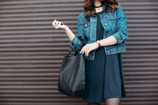 Crop of stylish incognito girl with big black leather bag after shopping, posing at street, leaning on wall. Woman in dress, jeans jacket, with accessories. Street fashion.