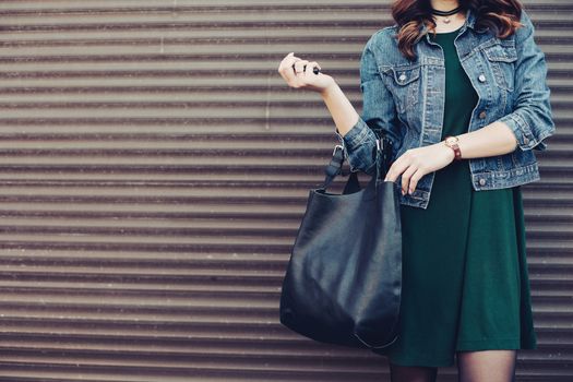 Crop of stylish incognito girl with big black leather bag after shopping, posing at street, leaning on wall. Woman in dress, jeans jacket, with accessories. Street fashion.