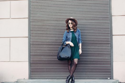 Stylish beautiful brunette woman in sunglasses, green dress, brown hat and big black bag, posing standing at street. Fashionable smiling girl leaning on wall. Concept of fashion and street trend look.