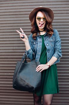 Stylish beautiful brunette woman in sunglasses, green dress, brown hat and big black bag, posing standing at street. Fashionable smiling girl leaning on wall. Concept of fashion and street trend look.