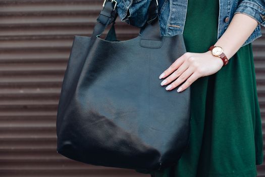 Crop of woman with big black leather bag after shopping, posing at street, showing perfect manicure, leaning on wall. Woman in dark green dress, jeans jacket, with accessories.
