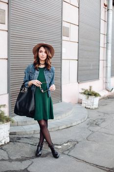 Fashionable brunette in brown hat, jeans jacket and choker on neck posing at street,looking at camera. Confident stylish woman with handbag, after beauty salon and shopping. Street swag style.