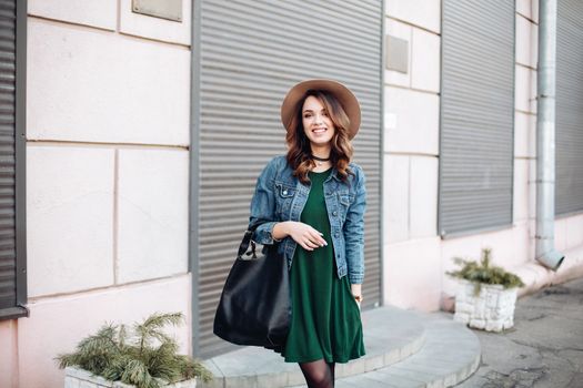 Stylish beautiful brunette woman in sunglasses, green dress, brown hat and big black bag, posing standing at street. Fashionable smiling girl leaning on wall. Concept of fashion and street trend look.