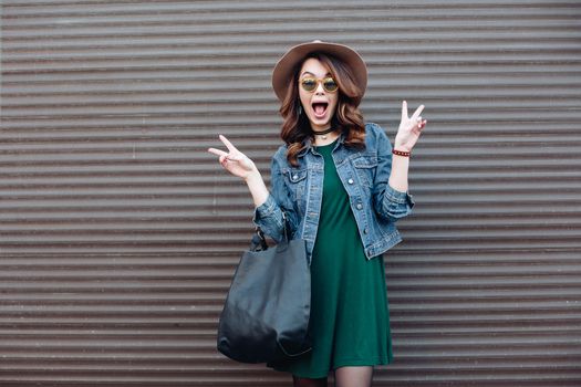 Stylish beautiful brunette woman in sunglasses, green dress, brown hat and big black bag, posing standing at street. Fashionable smiling girl leaning on wall. Concept of fashion and street trend look.