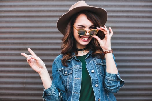 Fashion, beautiful brunette in brown hat, making duck face and showing peace by two hands. Attractive and stylish girl in sunglasses,choker, jeans jacket posing at street sending kiss at camera.