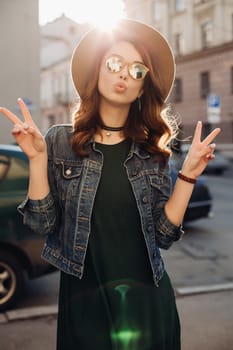 Fashion, beautiful brunette in brown hat, making duck face and showing peace by two hands. Attractive and stylish girl in sunglasses,choker, jeans jacket posing at street sending kiss at camera.