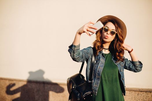 Brunette girl in sunglasses and hat making duck face, sending kiss at camera and taking self portrait at smart phone.Beautiful and funny woman posing, walking at street. Concept of street fashion.