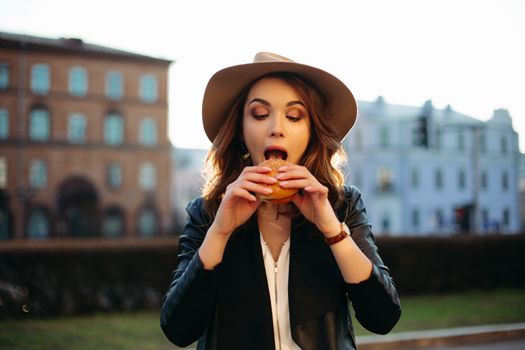 Funny portrait of stylish brunette girl in hat, posing with food, eating hamburger and surprised looking at camera. Emotionally beautiful woman in leather jacket, having dinner of fast food at street.
