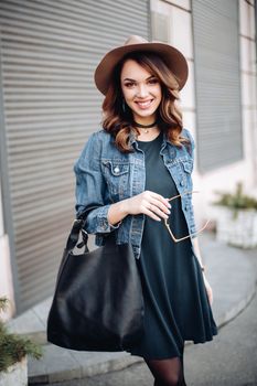 Fashionable brunette in brown hat, jeans jacket and choker on neck posing at street,looking at camera. Confident stylish woman with handbag, after beauty salon and shopping. Street swag style.