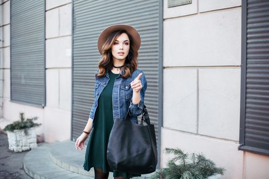 Stylish beautiful brunette woman in sunglasses, green dress, brown hat and big black bag, posing standing at street. Fashionable smiling girl leaning on wall. Concept of fashion and street trend look.