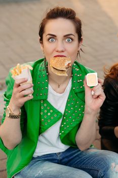 Funny girls sitting at street and eating fast food, having dinner together, and smiling to each other. Beautiful young girlfriends posing with hamburger and potato fried. Junk and unhealthy food.