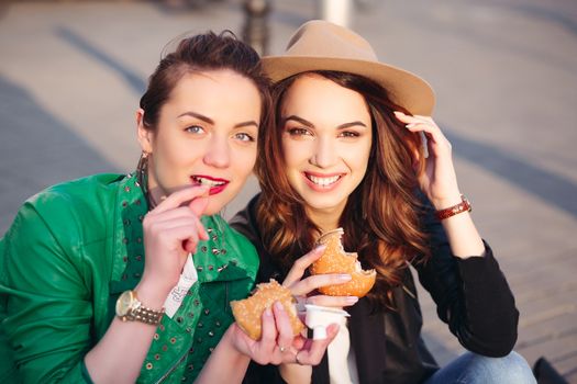 Funny girls sitting at street and eating fast food, having dinner together, and smiling to each other. Beautiful young girlfriends posing with hamburger and potato fried. Junk and unhealthy food.