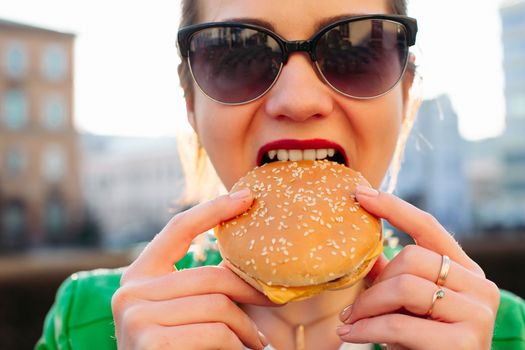 Beautiful and fashionable brunette woman in brown hat eating tasty hamburger with clossed eyes and enjoying. Emotionally girl having dinner of fast food at street, sensuality smiling.