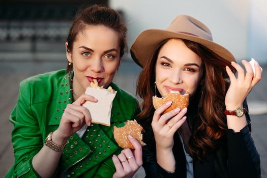 Funny girls sitting at street and eating fast food, having dinner together, and smiling to each other. Beautiful young girlfriends posing with hamburger and potato fried. Junk and unhealthy food.