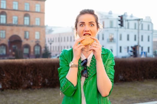 Beautiful and fashionable brunette woman in brown hat eating tasty hamburger with clossed eyes and enjoying. Emotionally girl having dinner of fast food at street, sensuality smiling.