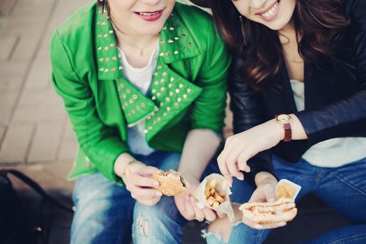 Funny girls sitting at street and eating fast food, having dinner together, and smiling to each other. Beautiful young girlfriends posing with hamburger and potato fried. Junk and unhealthy food.