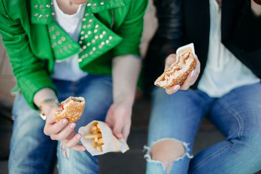 Funny girls sitting at street and eating fast food, having dinner together, and smiling to each other. Beautiful young girlfriends posing with hamburger and potato fried. Junk and unhealthy food.