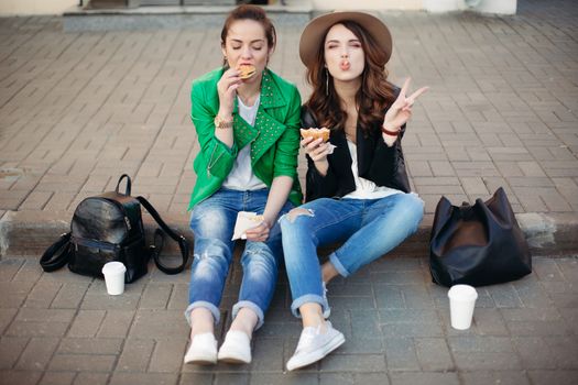 Funny girls sitting at street and eating fast food, having dinner together, and smiling to each other. Beautiful young girlfriends posing with hamburger and potato fried. Junk and unhealthy food.