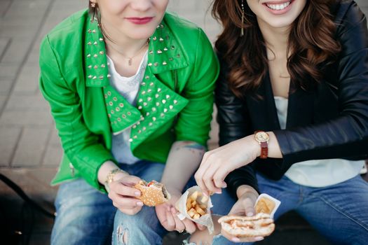 Funny girls sitting at street and eating fast food, having dinner together, and smiling to each other. Beautiful young girlfriends posing with hamburger and potato fried. Junk and unhealthy food.