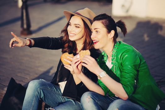 Two best friends two beautiful and stylish women, sitting at street, having dinner together, and telling each other gossip. Fashionable girls eating hamburgers and talking. Street style. Fast food.