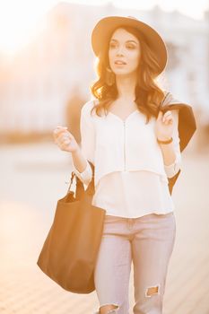 Beautiful positivity brunette woman in hat, white blouse, jeans walking at sunset in city, after shopping and beauty salon. Smiling fashionable girl stylish wearing posing at camera. Hipster look.