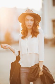 Beautiful positivity brunette woman in hat, white blouse, jeans walking at sunset in city, after shopping and beauty salon. Smiling fashionable girl stylish wearing posing at camera. Hipster look.