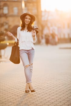 Beautiful positivity brunette woman in hat, white blouse, jeans walking at sunset in city, after shopping and beauty salon. Smiling fashionable girl stylish wearing posing at camera. Hipster look.