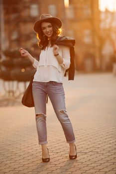 Beautiful positivity brunette woman in hat, white blouse, jeans walking at sunset in city, after shopping and beauty salon. Smiling fashionable girl stylish wearing posing at camera. Hipster look.