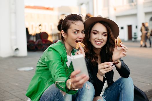 Positive and stylish girls eating hamburgers making duck face and taking self portrait at smart phone.Beautiful two women stylish wearing in hat and leather jacket, have dinner at street by fast food.