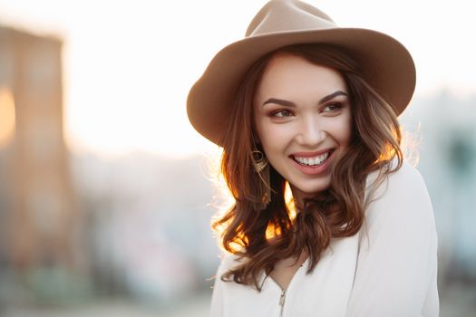 Portrait of young and beautiful brunette woman emotionally smiling, walking at street,posing, looking away. Girl in hat, with big earrings with perfect make up and wavy hairstyle. Street fashion look.