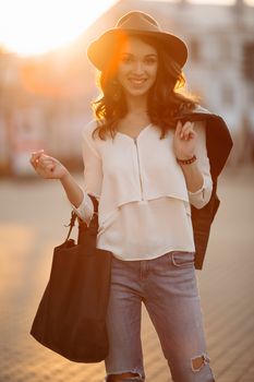 Beautiful positivity brunette woman in hat, white blouse, jeans walking at sunset in city, after shopping and beauty salon. Smiling fashionable girl stylish wearing posing at camera. Hipster look.