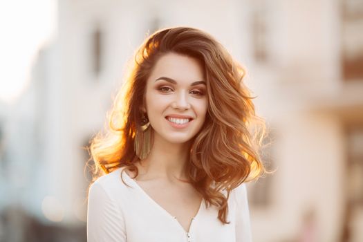 Portrait of young and beautiful brunette woman emotionally smiling, walking at street,posing, looking away. Girl in hat, with big earrings with perfect make up and wavy hairstyle. Street fashion look.