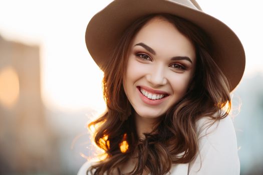 Portrait of young and beautiful brunette woman emotionally smiling, walking at street,posing, looking away. Girl in hat, with big earrings with perfect make up and wavy hairstyle. Street fashion look.