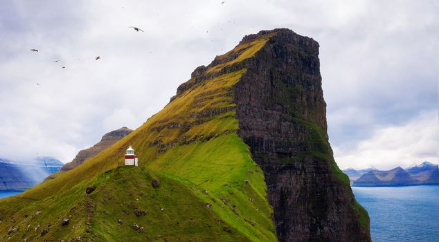 Small white Kallur lighthouse located near huge cliffs on island of Kalsoy with seagulls flying. Kalsoy is an isolated small island in the north-east of the Faroe Islands.