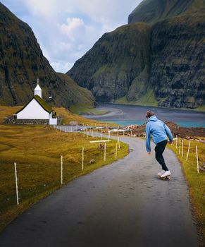 Young skater riding a skateboard on a road in the village of Saksun, Faroe Islands, surrounded by beatiful scenery