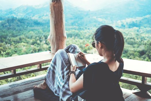 Woman is writing down in small white memo notebook for take a note plan to do or write a book with beautiful terrace of house and nature mountain view background.