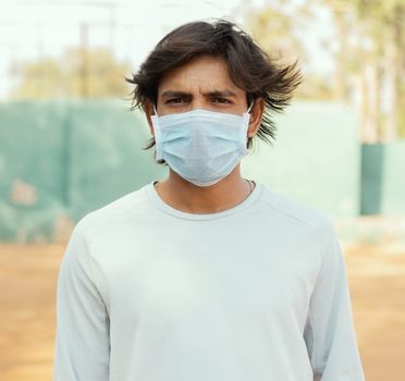 Young man with medical mask looking into the camera while wind blowing his hairs during hot sunny day - concept of wear mask outdoors to stop spreading and protect from covid-19 or coronavirus