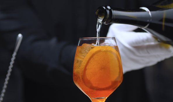 Preparing an Aperol Spritz Cocktail. Close up bartenders hands pouring prosecco in wine glass. Long fizzy drink.