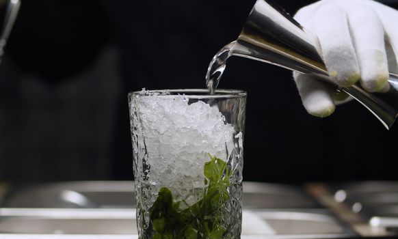 Close up making a Mojito Classic Cocktail. In a glass with mint and crushed ice, the bartender pours strong alcohol. Long drink.