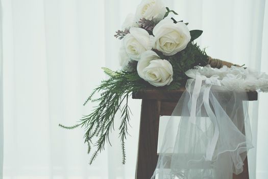 wedding white bridal veil & rose flower bouquet on wooden chair beside see through sheer window curtain