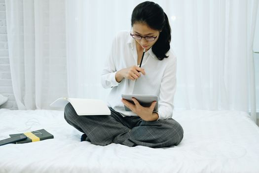 asian woman writing note watching video. student listen lecture study online course on bed in bedroom at home