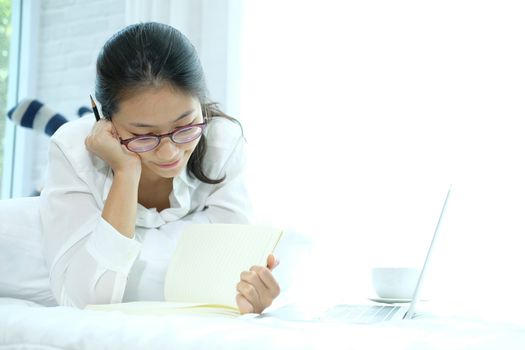 asian woman writing note working with computer laptop on bed. student listen lecture learning studying online in bedroom.