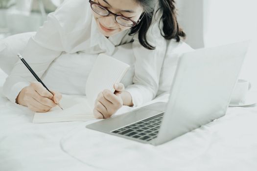 asian woman writing note working with computer laptop on bed. student listen lecture learning studying online in bedroom.