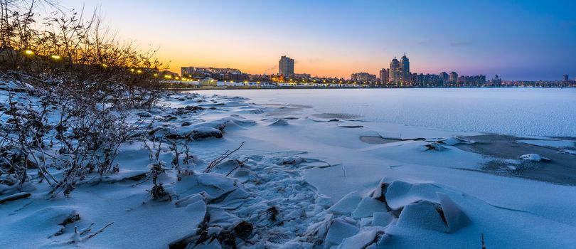 Cold Winter night cityscape with illuminated buildings in Kiev, Ukraine. The frozen Dnieper river appears in the foreground