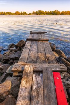 A wooden pontoon with a red paint plank  over big stones close to the blue Dnieper river is waiting for the fisherman.