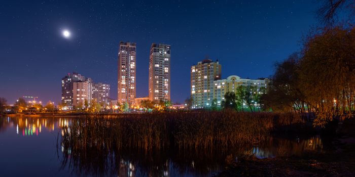 Night view of modern buildings in the obolon district of Kiev, Ukraine, viewed from the southern part of Natalka Park. The moon shines in the starry blue sky. The lights reflect on Dnieper river.