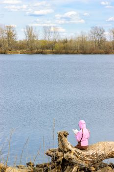 A woman dressed in pink, sitting on a tree trunk by the river is reading a book